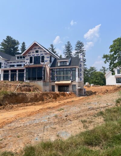 A large house under construction with visible scaffolding and building materials, situated on a dirt lot with some grass and trees in the background. A smaller completed white building is nearby.