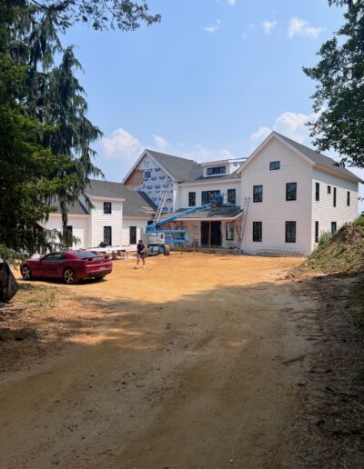 A large, white house under construction with scaffolding and building materials visible. A person stands near a red car parked on the dirt driveway in front of the house. Trees surround the area.