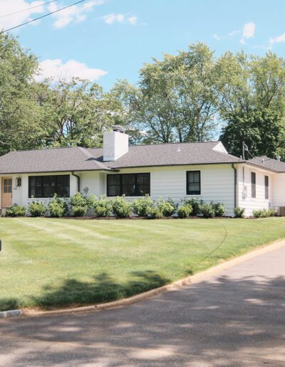 A single-story house with white siding and a black roof, surrounded by trees, features a neatly manicured lawn and a driveway with a black vehicle parked at the end. A black mailbox is in the foreground.