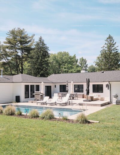 Single-story white house with a dark roof, outdoor seating area, and a swimming pool in the backyard, surrounded by a well-maintained lawn and tall trees.