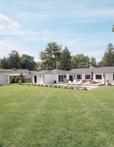 A single-story modern house with white exterior and dark roof, surrounded by a large grassy yard and trees. The backyard features a swimming pool and patio area.