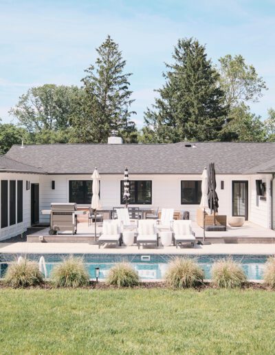 A white single-story house with a dark roof features a backyard pool, patio furniture, and a grill. Trees surround the property.