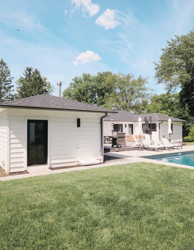 A backyard scene featuring a white house with black trim, a neatly manicured lawn, and an outdoor pool with sun loungers. Trees and shrubs border the yard under a partly cloudy sky.