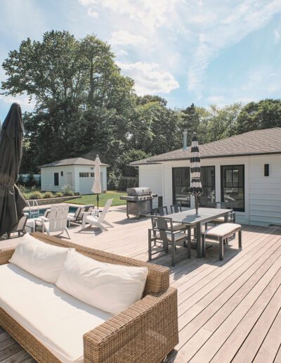 A spacious wooden deck featuring outdoor seating, a dining area, umbrellas, and a pool in the background, adjacent to a single-story house with large windows. Trees and a small building are in the yard.