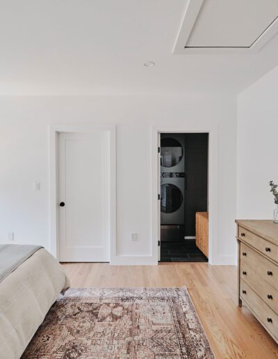 A modern bedroom with a bed, a dresser, a wall-mounted TV, and a door leading to a laundry area. The room features white walls, wooden floors, and a rug.