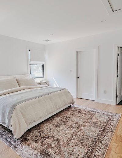 A minimalist bedroom with a white bed, wooden furniture, a large rug, and a laundry room visible through an open door. The room has light-colored walls and wooden flooring, with natural light from the windows.