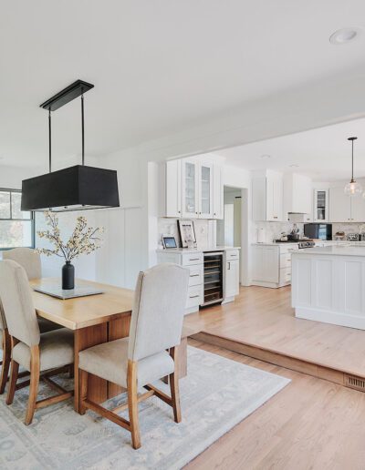 A modern open-concept dining area flows into a bright kitchen with an island and bar stools. The dining space features a wooden table with upholstered chairs and a large black pendant light above.