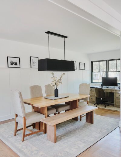 A well-lit dining area features a wooden table with chairs and a bench on a rug. A black rectangular pendant light hangs above the table. The space includes a built-in desk and white cabinetry.