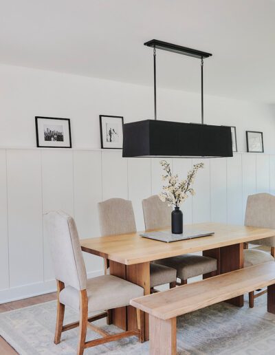 A modern dining room with a wooden table, four beige chairs, a bench, black rectangular light fixture, white walls, framed photos, and a desk with computer by the window.