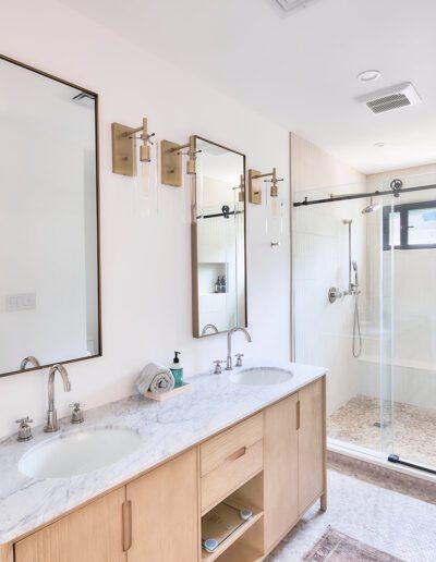 Modern bathroom with a double sink vanity on the left, marble countertop, two mirrors, and light fixtures above. A glass-enclosed shower is on the right with a bench and a window.