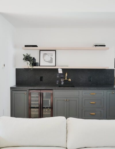 A modern kitchen area with dark gray cabinets, black countertops, a gold faucet, and minimal wall shelves. A white couch is in the foreground. A door and a partial view of a bathroom are in the background.