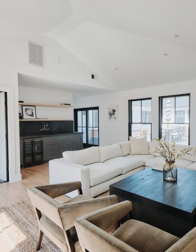 A modern living room with white walls, a beige sectional sofa, two brown chairs, a black coffee table, and large windows. A framed photograph and a door to a bathroom are visible in the background.