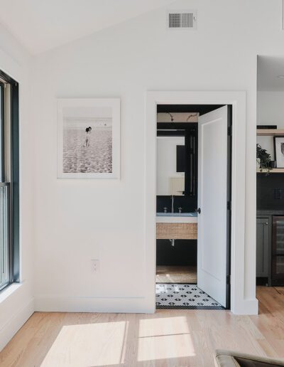 A contemporary living room with large windows, a closed door leading to a bathroom, dark cabinetry in the kitchen area, and a black-and-white photo on the wall. The room has light hardwood floors.