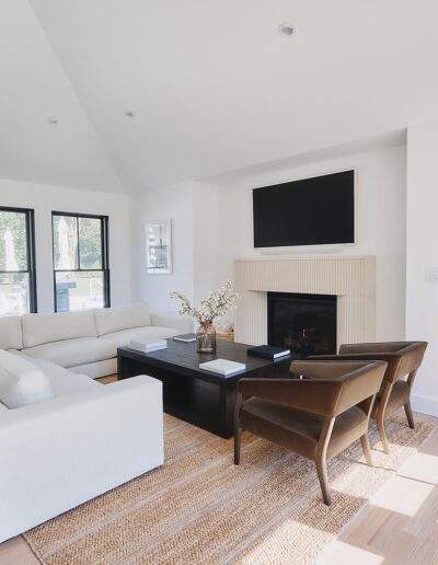 A modern living room featuring a white sectional sofa, two brown chairs, a black coffee table, a wall-mounted TV above a fireplace, and large windows with black frames. A hallway is visible in the background.