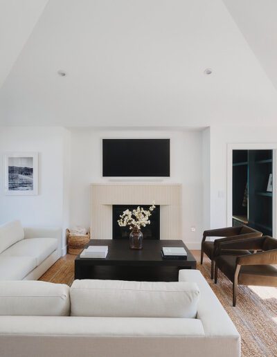 Modern living room with vaulted ceilings, a white sectional sofa, two dark brown chairs, a black coffee table, and a fireplace with a TV above it. Large windows provide ample natural light.