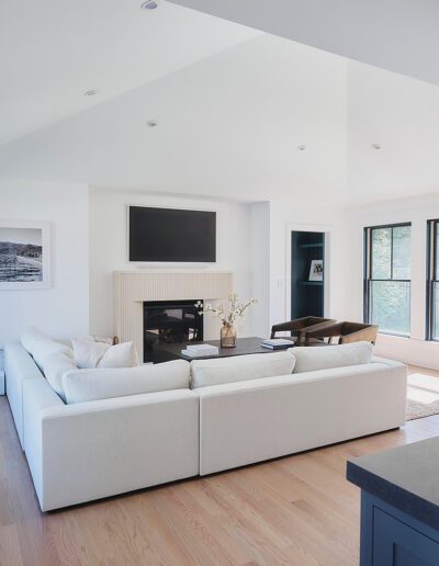 A modern living room features a large white sectional sofa, a fireplace with a TV above it, and windows letting in natural light. The space has light hardwood flooring and minimalist decor.