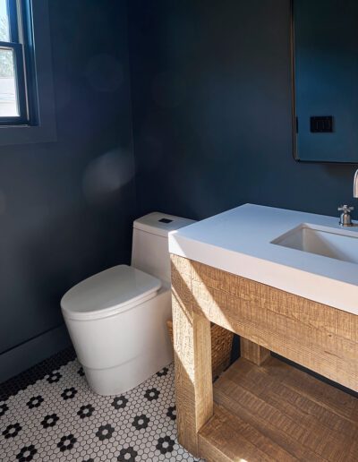 A small bathroom with dark walls, a modern toilet, and a white sink on a wooden vanity. The floor features black and white hexagonal tiles, and a window provides natural light.