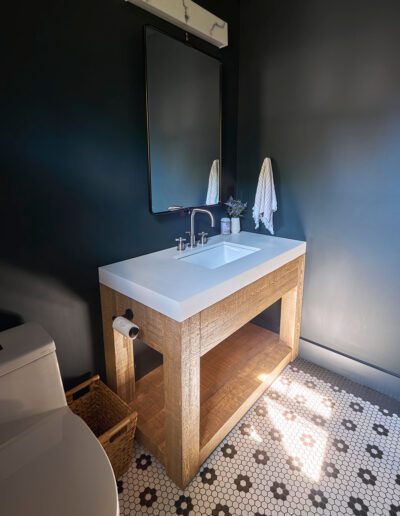 Bathroom with a wooden vanity featuring a white sink and faucet, a large mirror with a light fixture above, a towel hanging on the wall, a toilet to the left, and a patterned tile floor.