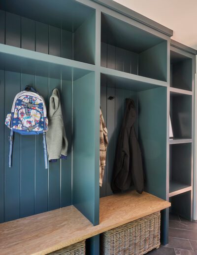 A mudroom with built-in green cubbies, hooks holding coats and a backpack, and handwoven baskets on wooden shelves beneath. Sunlight from windows brightens the interior.