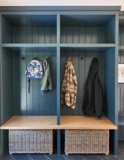 A blue entryway storage unit with cubbies, two hanging jackets, a backpack, two baskets underneath, and shelves on the right. There is a window on the left and a photo frame on the shelf.