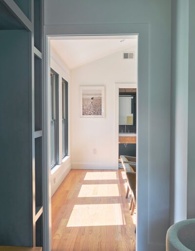 A hallway with a dark coat hanging on the left leads to a sunlit room with wooden flooring, a desk, a chair, and a framed photograph on the wall.