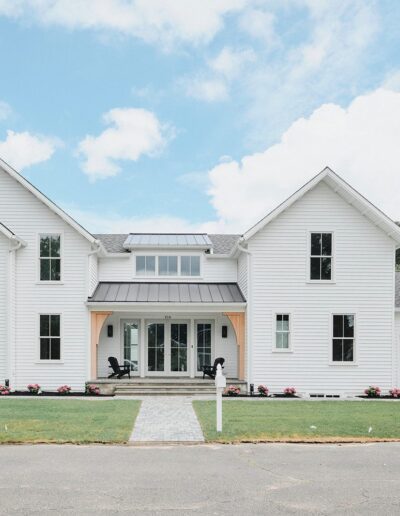 A large, modern white house with a symmetrical design, many windows, a gray roof, and a front yard with a pathway, lawn, and flower beds.