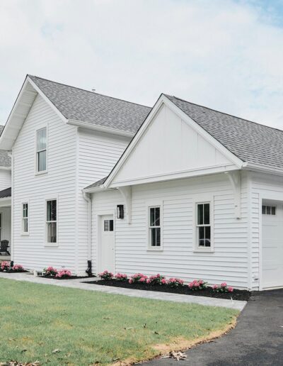 A white, two-story house with a large attached garage and a manicured front lawn. The house features multiple windows and a grey shingled roof, with a flower bed lining the front.