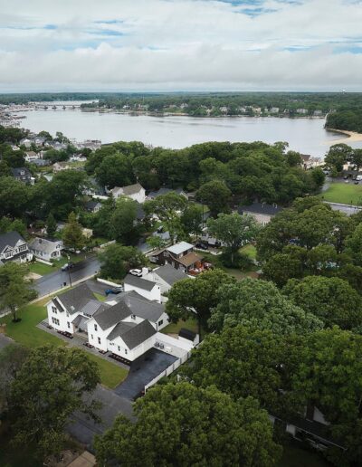 Aerial view of a residential neighborhood with numerous houses surrounded by dense trees, adjacent to a river with a sandy shoreline in the background under a partly cloudy sky.