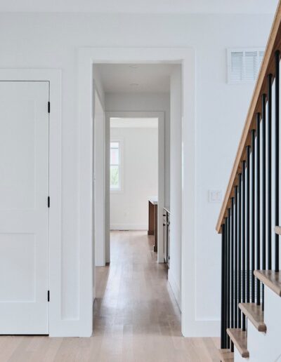 A hallway with light wood floors, white walls, a closed double door on the left, a staircase with black railings on the right, and an open doorway leading to another room.