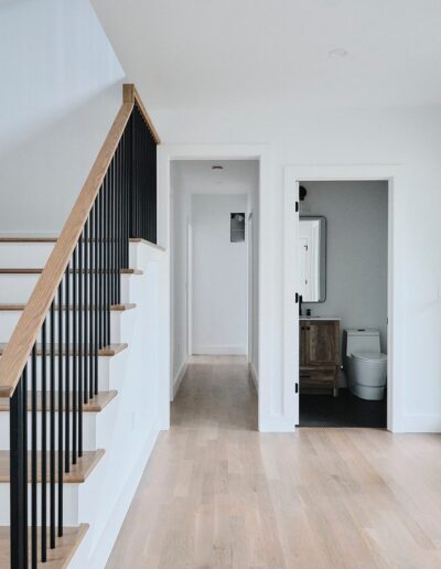 A modern, minimalistic hallway featuring light wood floors, white walls, a staircase with wooden steps and black railings, as well as a doorway leading to a bathroom.