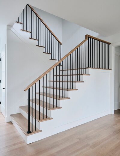 A modern wooden staircase with black metal railings is shown in a bright room with white walls and light wood flooring.