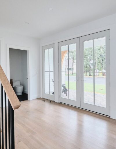 Bright hallway with light wooden floors, a wooden staircase with black railings, and glass doors leading to a patio. A small room with a white toilet is seen to the left.