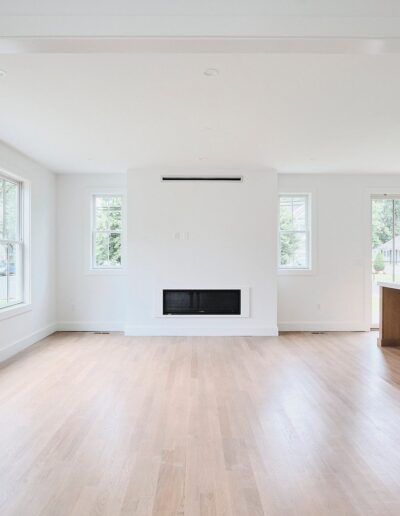 A spacious, unfurnished living room with light wood flooring, large windows, a built-in fireplace, and a view of an attached kitchen area.