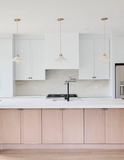 Modern kitchen with white cabinets, marble backsplash, stainless steel appliances, and a large wooden island with a white countertop. Three hanging pendant lights are above the island. Large windows on the left.