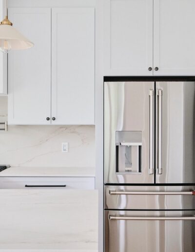 A modern kitchen with white cabinetry, a stainless steel refrigerator, a gas stove, and a light fixture hanging above a marble countertop.