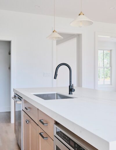 Modern kitchen with a quartz countertop island, built-in sink, black faucet, wooden cabinets, and stainless steel appliances. Two pendant lights hang overhead, with a window and door in the background.