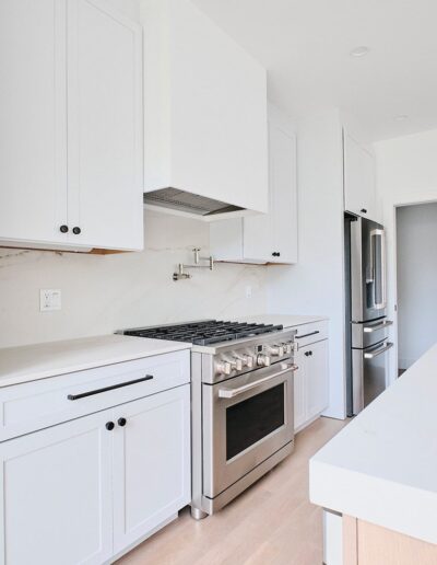 Modern kitchen with white cabinetry, stainless steel appliances, a gas stove, and a large island with a sink. The kitchen features minimalist design with built-in shelves and light wood flooring.