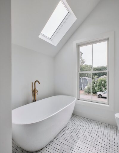 A modern bathroom featuring a white freestanding bathtub under a skylight and adjacent to a window. The floor is tiled with a small black and white mosaic pattern.