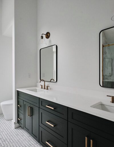 A modern bathroom with two mirrors above a long double-sink vanity, black cabinetry, brass fixtures, a window, and a glass-enclosed shower.