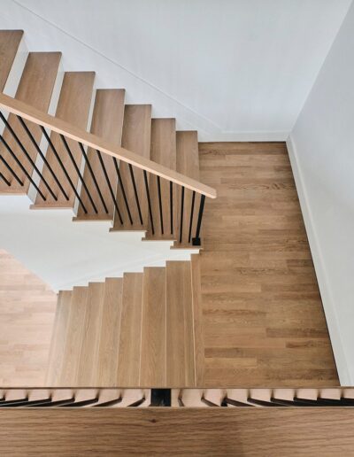 A bird's-eye view of a modern wooden staircase with black metal railings descending in a zigzag pattern, surrounded by white walls and hardwood floors.