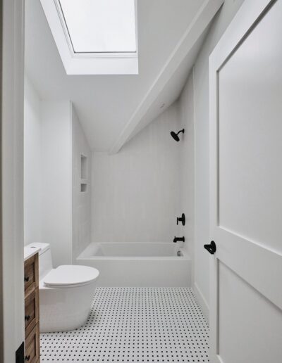 A modern bathroom with white walls, a skylight, a bathtub with a shower, a toilet, a wooden vanity, and a black and white checkered floor.