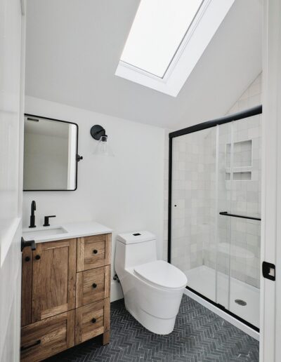 A modern bathroom with a wooden vanity, white countertop, rectangular mirror, toilet, glass-enclosed shower, and a skylight above. Black fixtures and herringbone floor tiles complete the design.