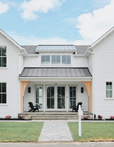 A white, two-story house with a large front porch, black roof accents, and manicured lawn. Two black Adirondack chairs are placed on the porch, facing a paved pathway leading to the entrance.